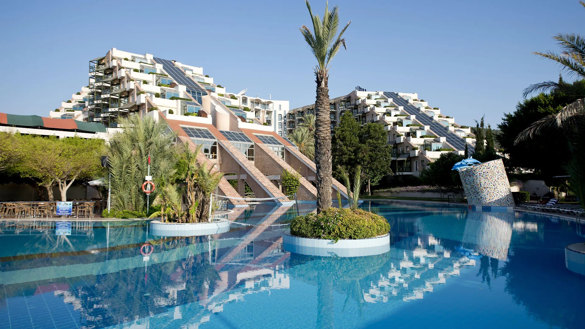 Limak Limra Hotel & Resort Kemer - Kids Concept Skyline photo The photo shows a vibrant resort scene on a sunny day. The foreground is dominated by a large, clear blue swimming pool, reflecting the sky and the surrounding buildings like a mirror. 

Several palm trees are scattered throughout the pool area, addi