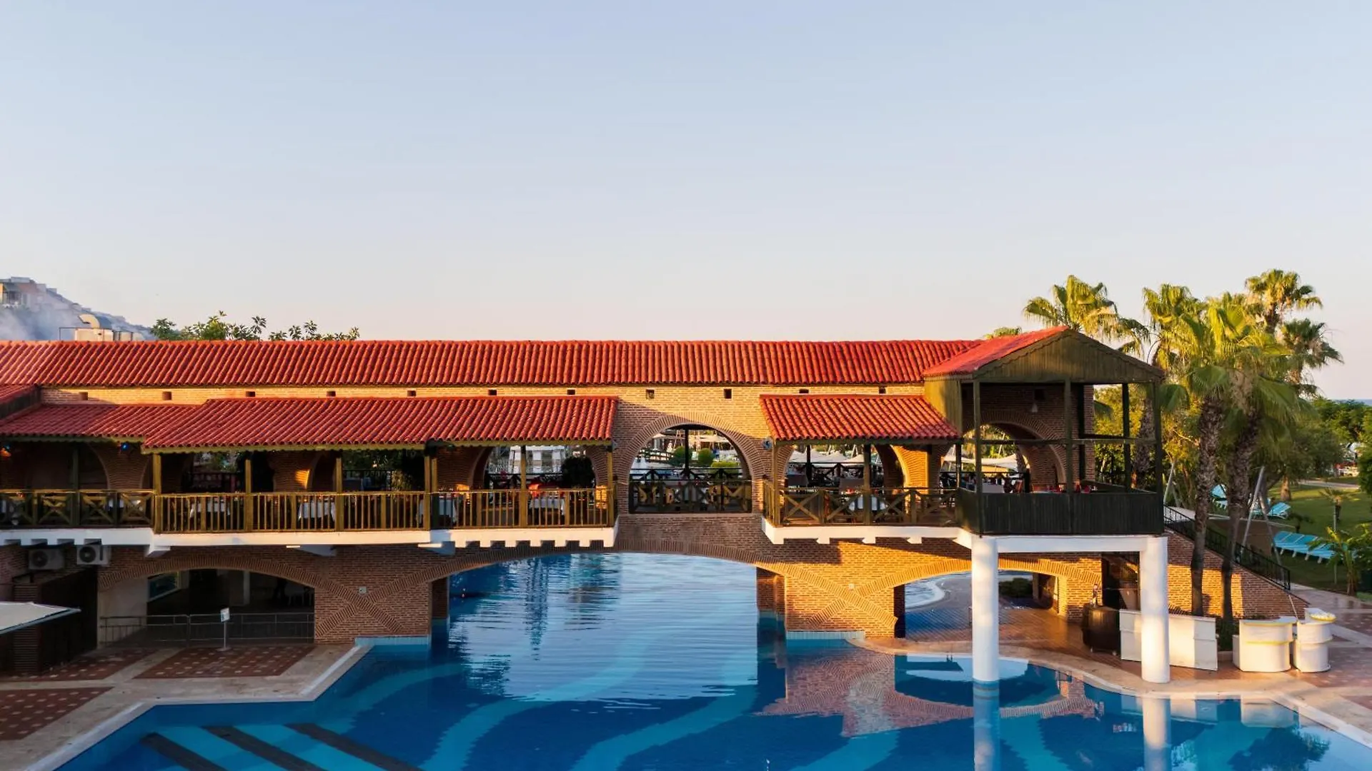 Limak Limra Hotel & Resort Kemer - Kids Concept Exterior photo The photo shows a beautiful resort or hotel complex centered around a large, inviting swimming pool. The pool has a vibrant blue color with wavy patterns created by blue and brown tiles.  

The pool is surrounded by a paved area and elevated walkways